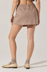 A person is wearing a high-waisted, taupe-colored suede mini skirt with a simple, clean design. They are also wearing beige loafers and light-colored socks. The image focuses on the back view, showcasing the skirt's smooth texture and minimalist style.