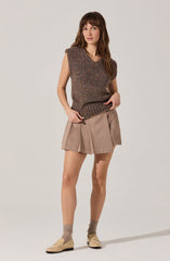 A woman is wearing a sleeveless, dark-colored knit vest with multicolored specks, paired with beige pleated shorts. She has on beige loafers and light brown socks.