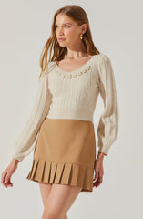 A woman is wearing a cream-colored, cable-knit sweater with long sleeves and a round neckline decorated with a pearl embellishment. She is also wearing a tan mini skirt with a pleated hem.