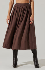 A woman is wearing a high-waisted, full-length brown skirt with pleats, paired with tall black boots.