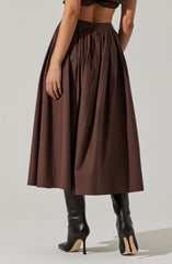 A woman is wearing a high-waisted, floor-length, dark brown skirt with a pleated design. The skirt has a zipper closure at the back. She is also wearing black high-heeled boots that reach just below the knees, with a pointed toe and stiletto heel.