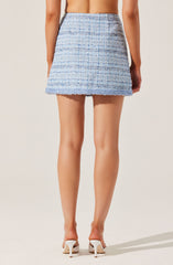 A woman is wearing a high-waisted, A-line mini skirt made of a textured, woven fabric with a pattern of light blue, white, and gray horizontal stripes. The skirt has a slight frayed hem at the bottom. She is also wearing white high-heeled shoes. The image shows her from the back, with her arms relaxed by her sides.