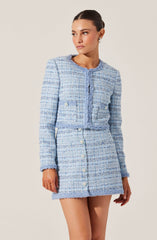 A woman is wearing a matching light blue tweed jacket and skirt set. The jacket is cropped with long sleeves, a round neckline, and decorative buttons on the front and on the sides. The skirt is high-waisted with a row of buttons down the front. The fabric has a textured, woven appearance with a mix of light blue, white, and darker blue threads. The woman has dark hair pulled back and is accessorized with hoop earrings.