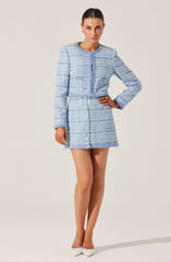 A woman is wearing a light blue, textured tweed skirt suit with matching jacket. The jacket has a round neckline, long sleeves, and a row of buttons down the front. The skirt is high-waisted with a row of buttons down the front. She is also wearing white pointed-toe heels.