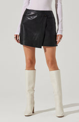 A person is wearing a black leather mini skirt with a single button closure and a slit on the side. They are also wearing a long-sleeved, light-colored top and tall, white high-heeled boots.