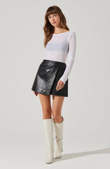 A woman is wearing a long-sleeved, semi-sheer white sweater with subtle horizontal stripes, paired with a black leather mini skirt featuring an asymmetrical wrap design and a side zipper. She completes her outfit with tall, off-white knee-high boots.