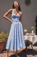 A woman is wearing a light blue sleeveless dress with wide straps and a fitted bodice. The dress features a pattern of evenly spaced brown polka dots. The skirt of the dress is full and long, reaching down to her ankles.