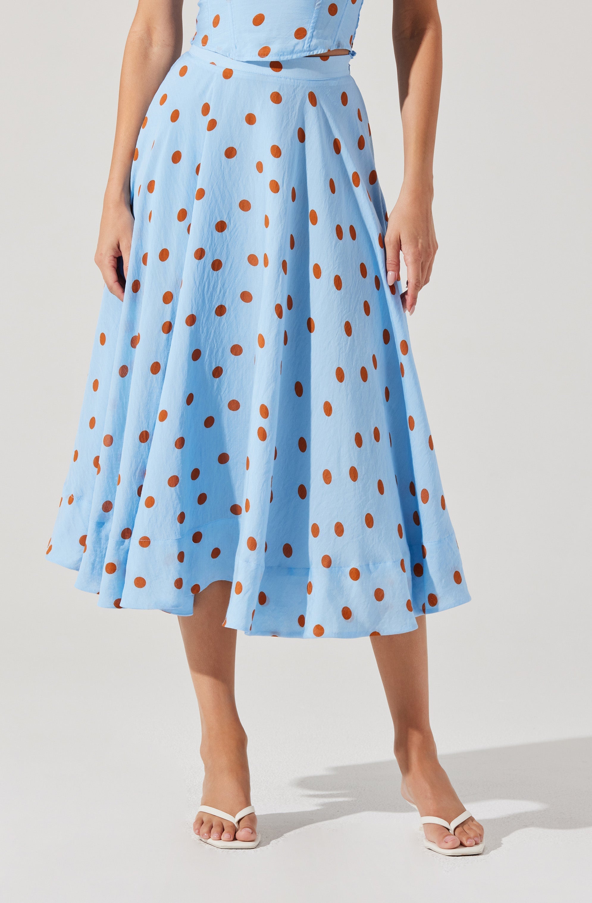 A woman is wearing a light blue, ankle-length skirt with an orange polka dot pattern. The skirt has a high waist and pleats, creating a full, flowing silhouette. She is also wearing a matching light blue top with the same orange polka dots.
