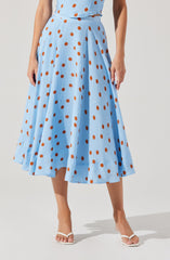A woman is wearing a light blue, ankle-length skirt with an orange polka dot pattern. The skirt has a high waist and pleats, creating a full, flowing silhouette. She is also wearing a matching light blue top with the same orange polka dots.