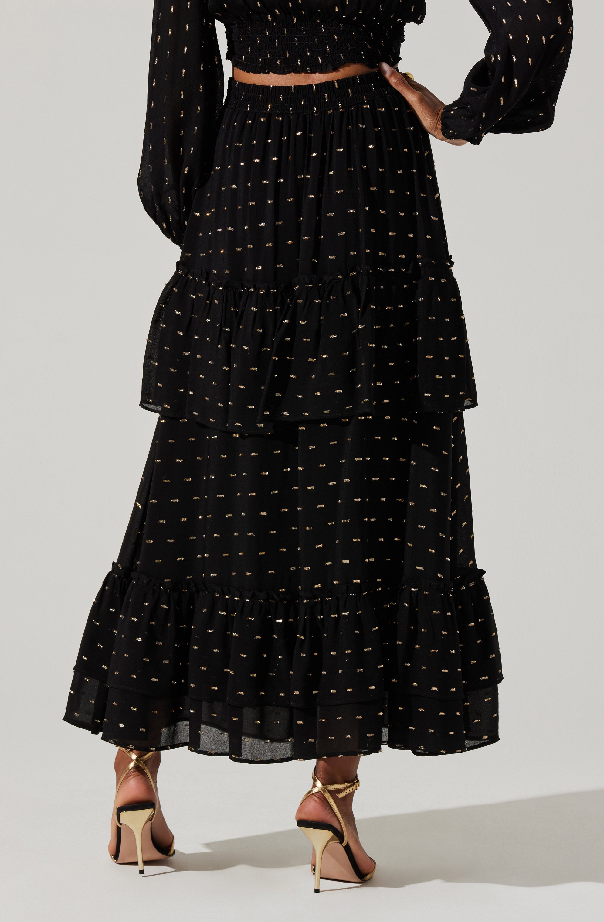 A woman is wearing a black, long, tiered skirt with small, evenly spaced gold dots. The skirt has ruffled tiers at the bottom. She is also wearing a matching black long-sleeved blouse with similar small gold dot patterns, with the sleeves slightly puffed.