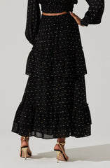 A woman is wearing a black, long, tiered skirt with small, evenly spaced gold dots. The skirt has ruffled tiers at the bottom. She is also wearing a matching black long-sleeved blouse with similar small gold dot patterns, with the sleeves slightly puffed.