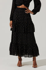 A woman is wearing a long black skirt with gold dotted patterns and ruffled tiered sections. She is also wearing a matching black top with gold dotted patterns, which is tied in a knot at the waist, revealing her midriff. The outfit is complemented by gold high-heeled sandals.