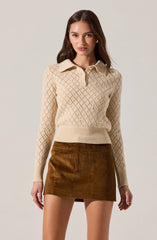 A woman is wearing a long-sleeved, cream-colored knit sweater with a diamond pattern. The sweater has a collar and a button-up neckline. She is also wearing a high-waisted, brown corduroy mini skirt with a front pocket.