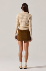 A person is wearing a cream-colored, long-sleeved sweater with a diamond-knit pattern, paired with a brown corduroy skirt. The individual is standing with their back to the camera, showing the sweater's fitted waistband and the skirt's above-the-knee length. They are also wearing beige shoes with low heels and cream-colored socks.