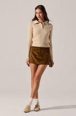 A woman is wearing a cream-colored, long-sleeved, knit sweater with a diamond pattern and a collar with buttons. She pairs it with a brown suede mini skirt and beige socks. She is also wearing beige slip-on shoes.