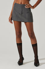 A woman is wearing a gray plaid mini skirt with a flat waistband and a small back pocket detail. She pairs this with sheer black knee-high socks and black high-heeled shoes with pointed toes and decorative bows.