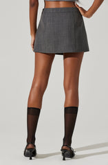 A woman is wearing a high-waisted, gray plaid mini skirt with a subtle checkered pattern, paired with black sheer knee-high socks and black high-heeled shoes with ankle straps. She is also wearing a cropped top that exposes her midriff.