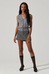 A woman is wearing a sleeveless, button-up, V-neck gray vest with a ribbed hem. She has on a high-waisted, dark gray plaid mini skirt. She is also wearing black sheer knee-high socks and black pointed-toe heels with bows.