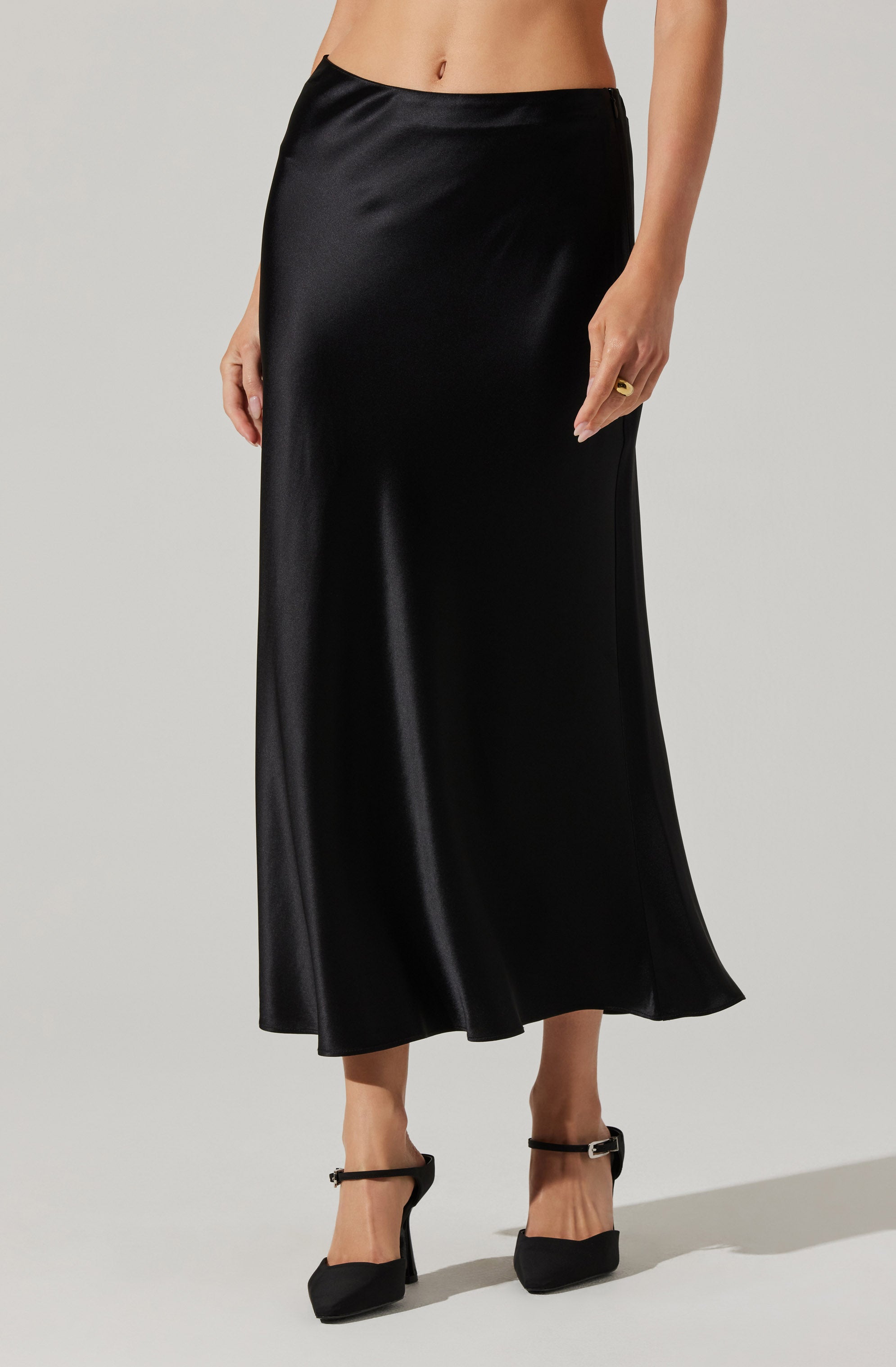 A woman is wearing a long, black satin skirt that reaches her ankles. She has a ring on her right hand and is wearing black high-heeled shoes with ankle straps.