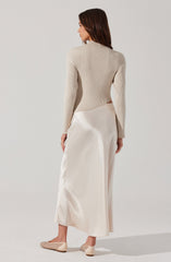 A woman is wearing a beige, long-sleeved, ribbed knit sweater with a high neckline. She is also dressed in an elegant, floor-length, satin-like skirt in a creamy off-white color. Her footwear consists of beige, low-heeled, woven-style shoes.