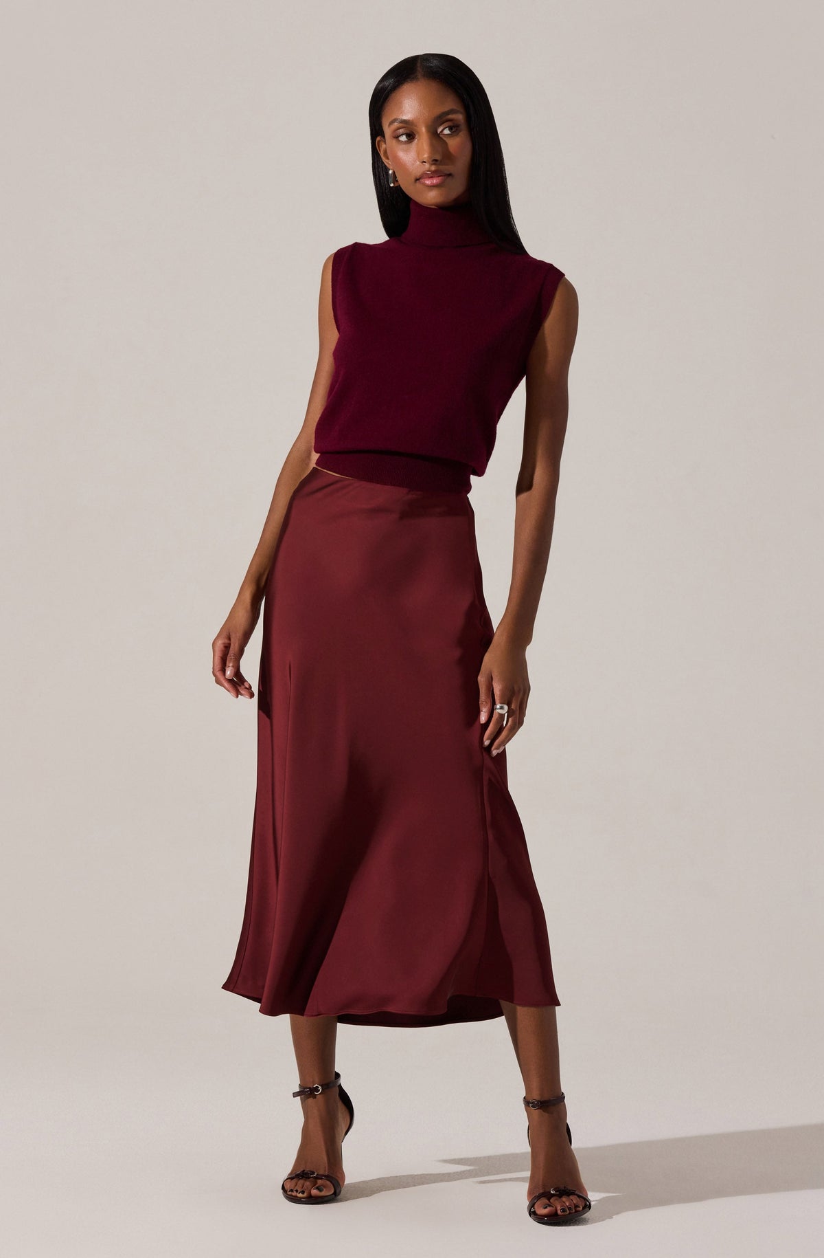 A woman is wearing a sleeveless, burgundy turtleneck top and a matching satin midi skirt with a slight slit on one side. She is also wearing black high-heeled sandals with ankle straps.