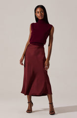 A woman is wearing a sleeveless, burgundy turtleneck top and a matching satin midi skirt with a slight slit on one side. She is also wearing black high-heeled sandals with ankle straps.