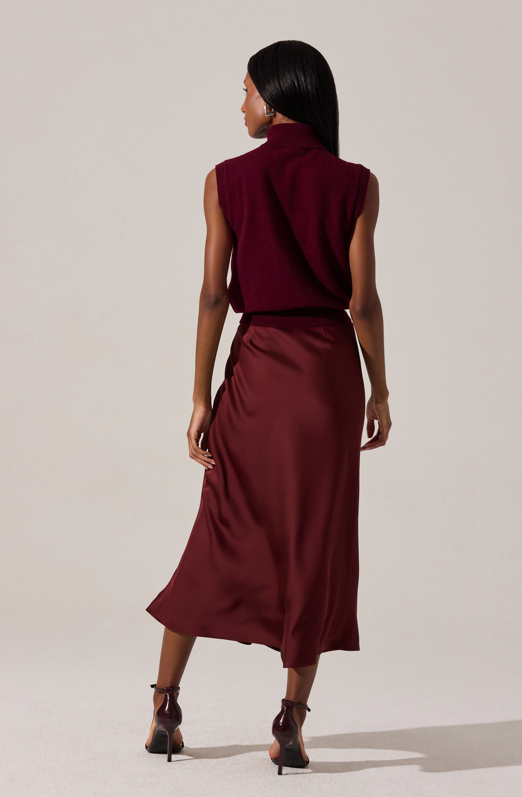 A woman is wearing a sleeveless, high-neck burgundy top with shoulder padding, paired with a matching satin midi skirt that has a slight sheen and a slit at the back. She is also wearing dark high-heeled shoes with ankle straps.