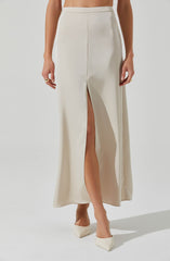 A woman is wearing a high-waisted, full-length cream-colored skirt with a front slit revealing her left leg. She has a ring on her right hand and is wearing cream-colored pointed-toe heels.