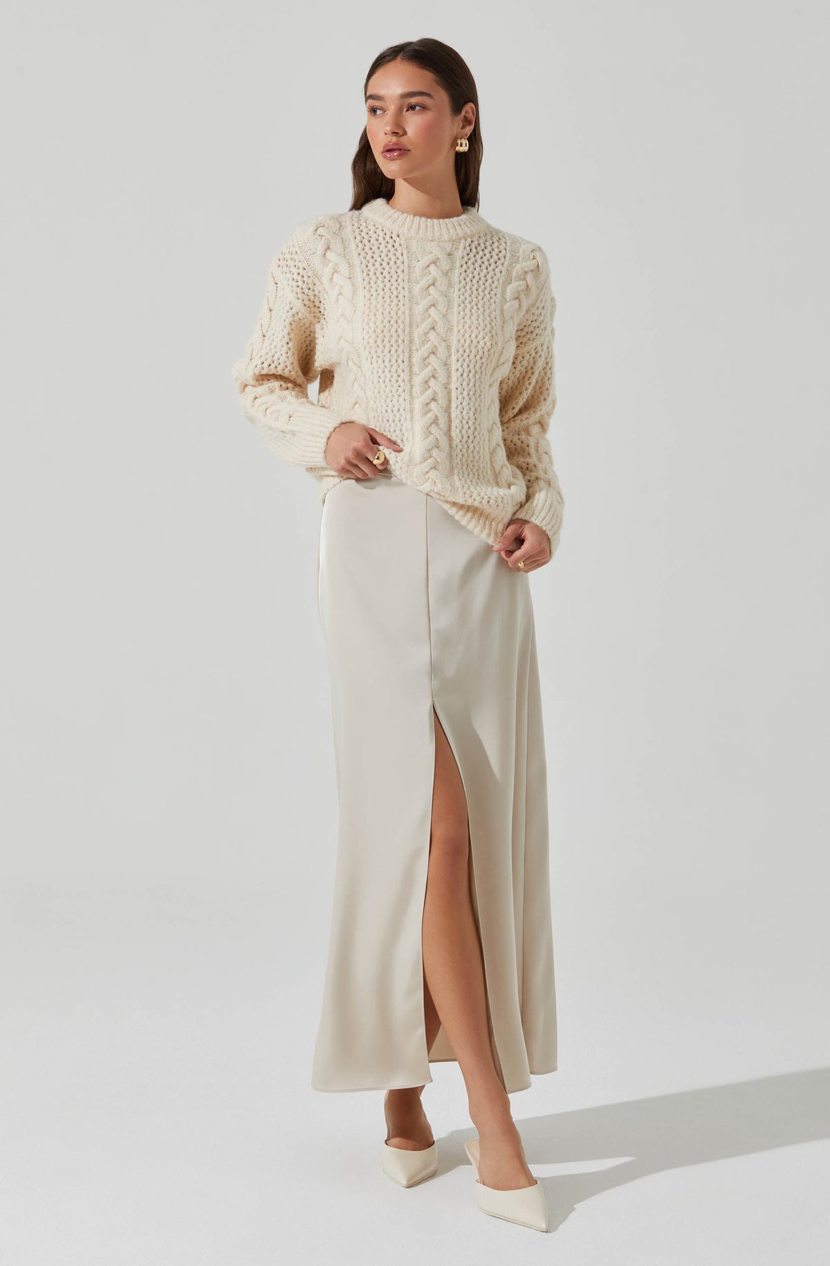 A woman is wearing an oversized cream-colored cable-knit sweater with a textured pattern on the front and long sleeves. She pairs it with a high-waisted, cream-colored, floor-length skirt that features a front slit revealing her leg. She completes the look with matching cream-colored heels and gold hoop earrings.