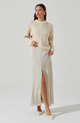 A woman is wearing an oversized cream-colored cable-knit sweater with a textured pattern on the front and long sleeves. She pairs it with a high-waisted, cream-colored, floor-length skirt that features a front slit revealing her leg. She completes the look with matching cream-colored heels and gold hoop earrings.
