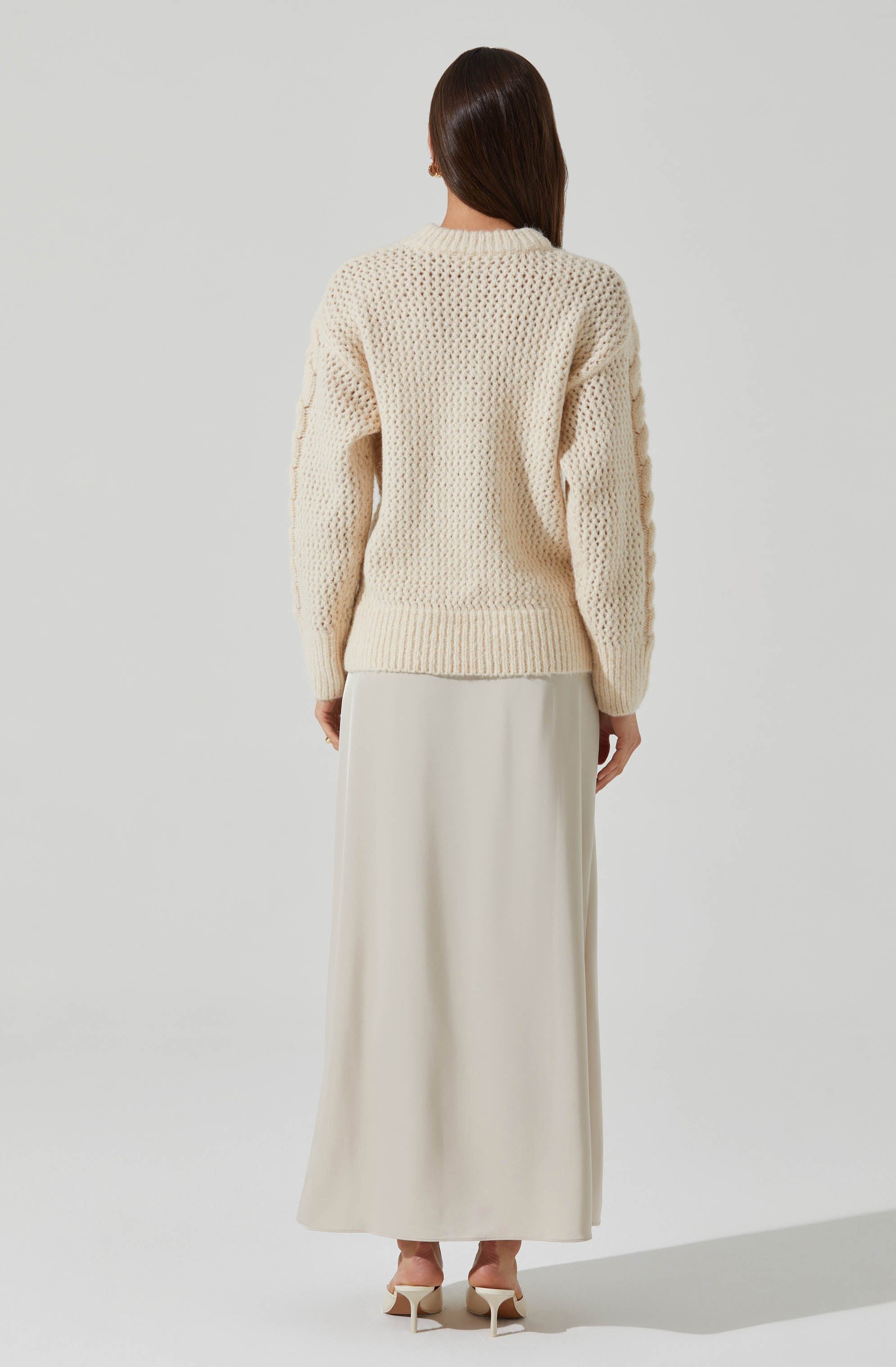 A woman is wearing an oversized, cream-colored, chunky knit sweater with a textured pattern. She pairs it with a long, flowing, cream-colored skirt and nude high-heeled shoes. The background is plain and light-colored.