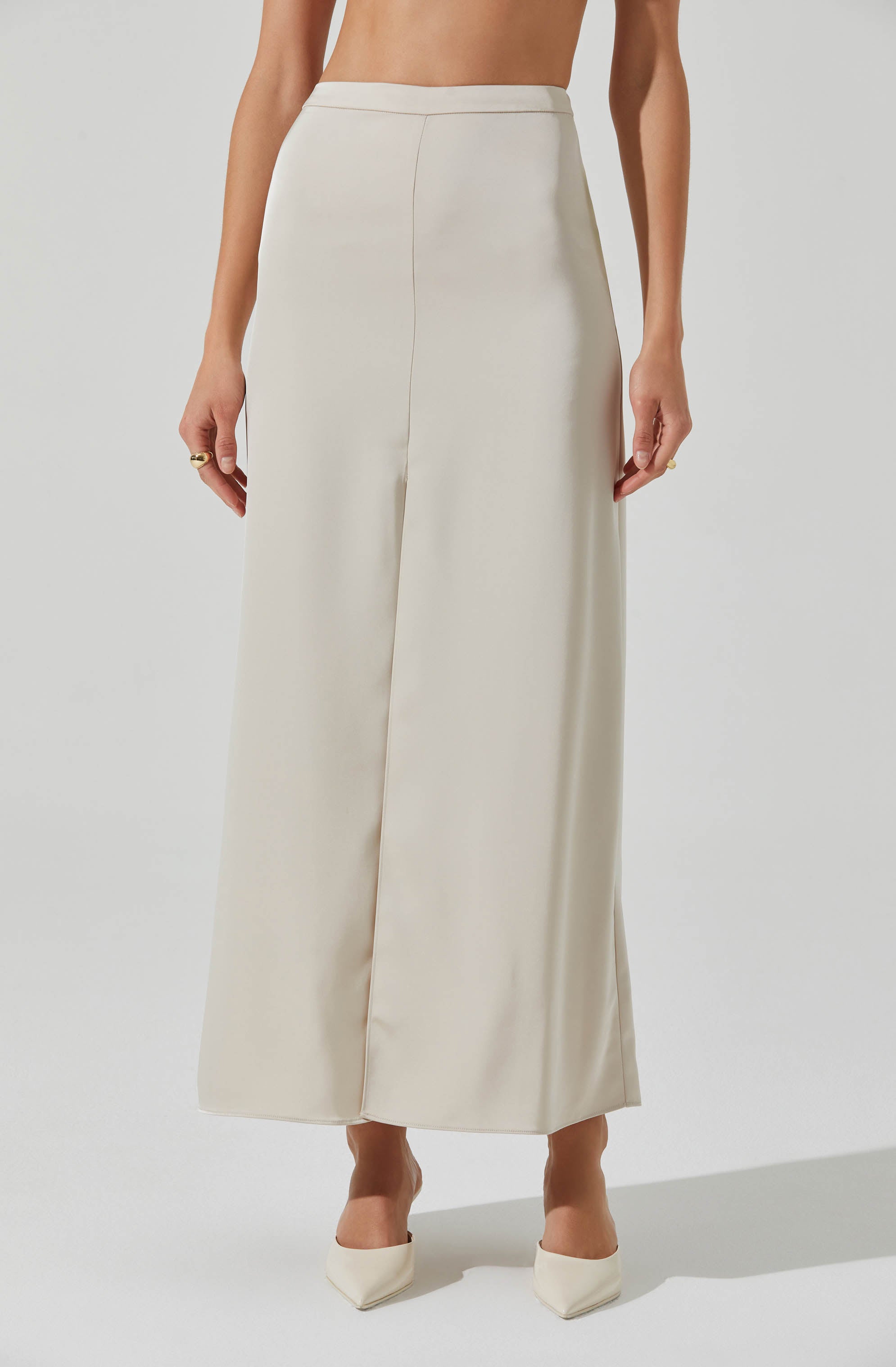 A person is wearing high-waisted, wide-leg cream-colored trousers with a smooth, silky fabric. The trousers have a clean, tailored waistband and a front center seam running from the waistband to the hem. The person is also wearing matching cream-colored pointed-toe shoes.