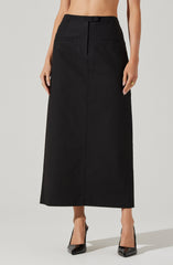 A model wearing a long, high-waisted black skirt with a straight silhouette, featuring a front button closure, belt loops, and two horizontal slit pockets on the front. The skirt extends down to the ankles, paired with black pointed-toe high heels.