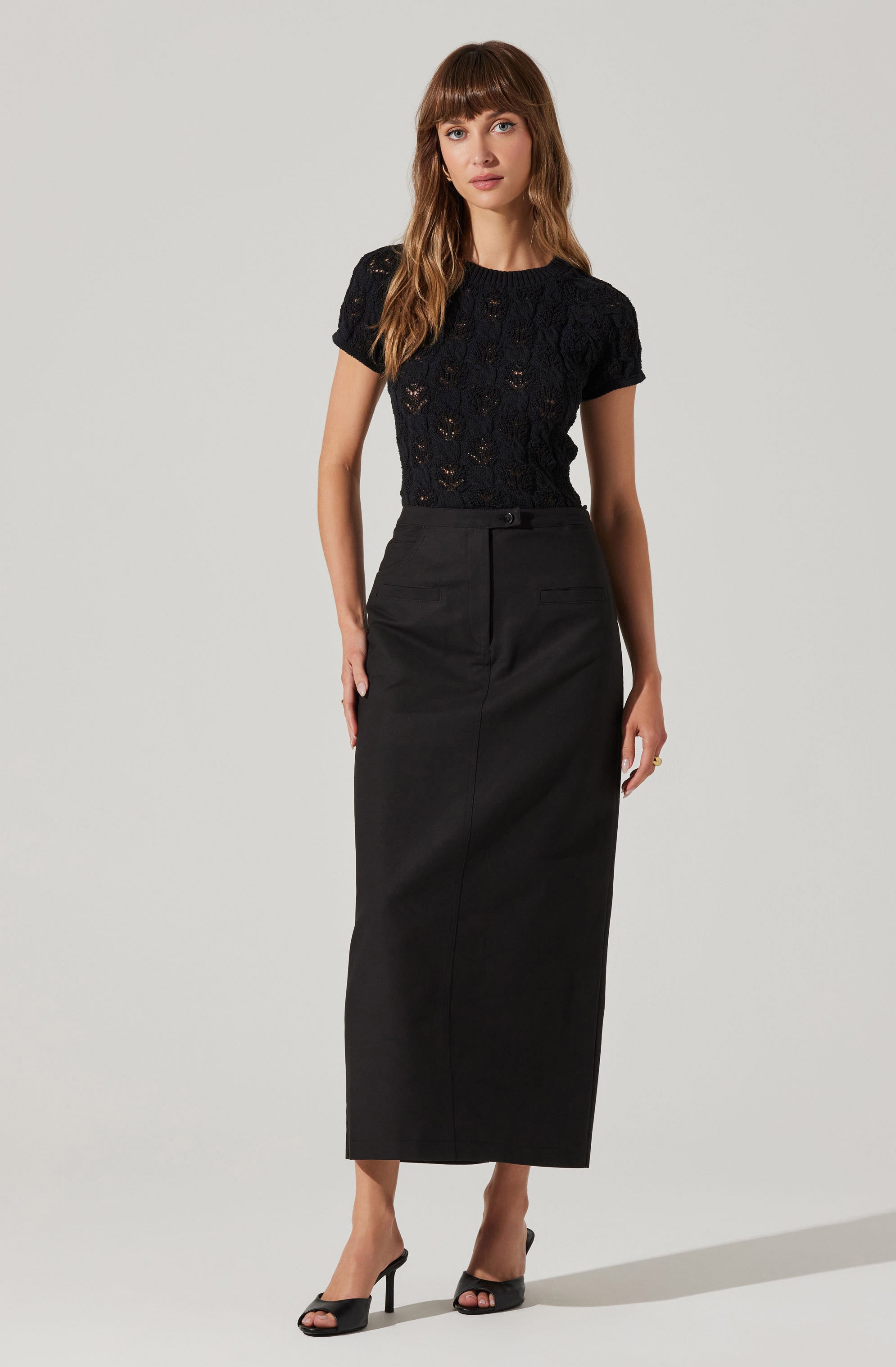 A woman is wearing a short-sleeved black lace top with intricate floral patterns, paired with a high-waisted, ankle-length black skirt that features a front button and hidden zipper. She completes her outfit with black open-toe high-heeled sandals.