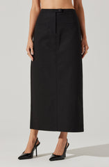 A woman is wearing a high-waisted, full-length black skirt with a straight fit and a button and zipper closure at the waistband. The skirt has two small, horizontal front pockets. She is also wearing black high-heeled pointed-toe shoes.