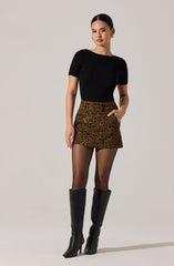 A woman is wearing a fitted black short-sleeve ribbed top, a high-waisted leopard print mini skirt, sheer black tights, and tall black boots with a high heel. She has her hair slicked back and is accessorized with large gold hoop earrings and a gold ring.