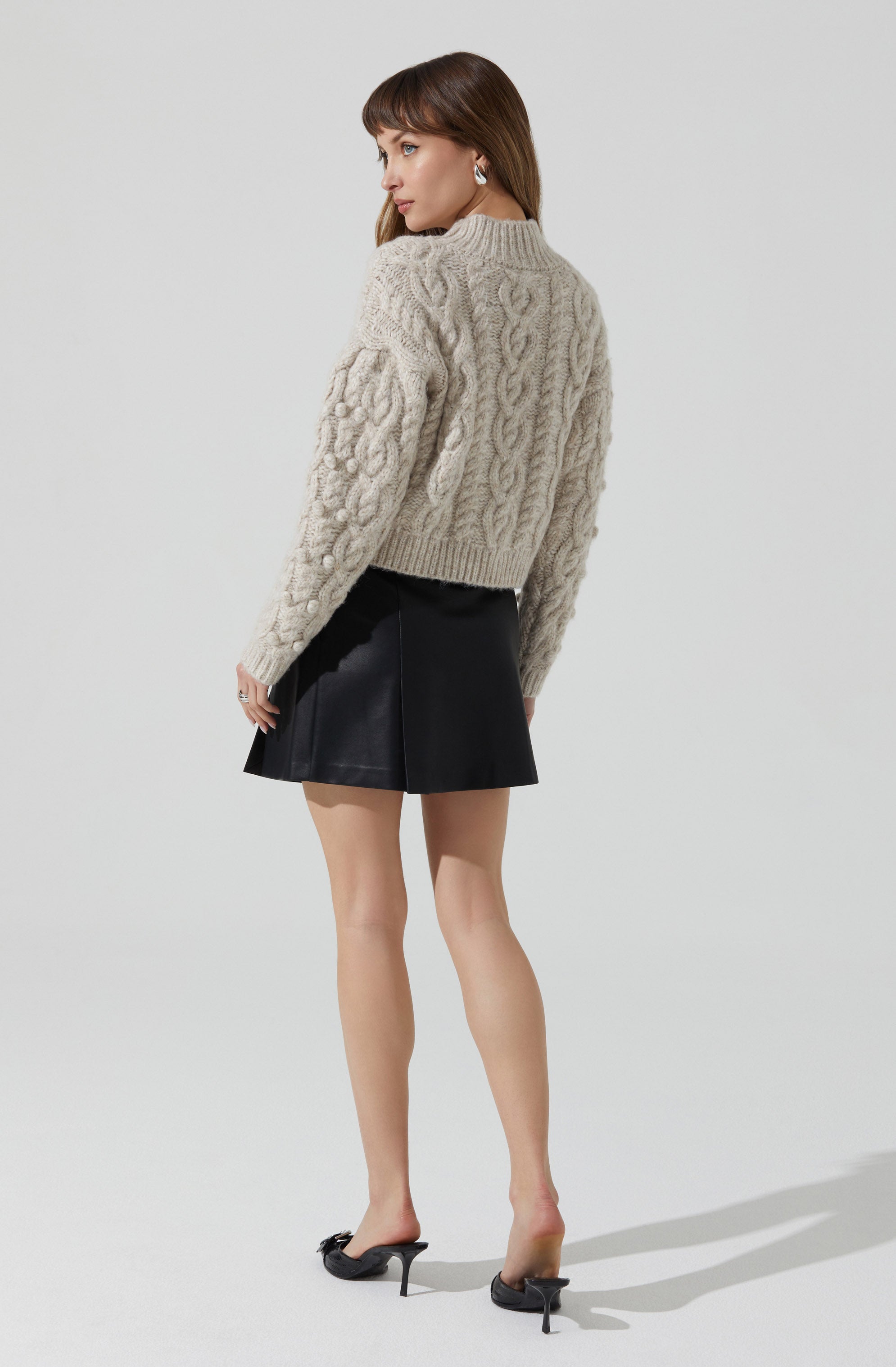 A woman is wearing a chunky, beige knit sweater with cable and bobble patterns, paired with a black leather A-line mini skirt. She is also wearing black high-heeled mules.