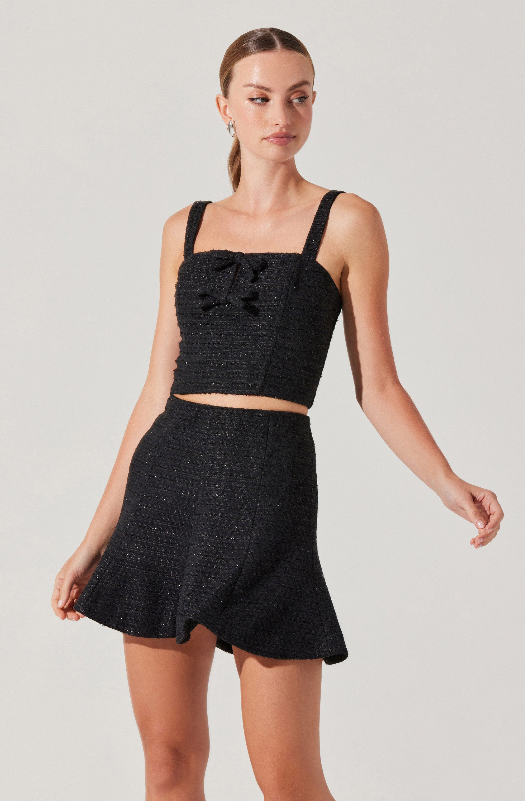 A woman is wearing a matching black set consisting of a sleeveless cropped top with wide straps and a bow detail at the front, paired with high-waisted, flared shorts. The fabric appears to have a textured, slightly shiny finish.
