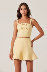 A young woman is wearing a light yellow two-piece outfit consisting of a sleeveless crop top with thick straps and a decorative bow at the chest, paired with a matching high-waisted, flared skirt. The fabric of both pieces has a subtle textured pattern.