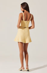 A woman is wearing a sleeveless, light yellow outfit with a fitted top that has wide straps and a zipper closure in the back, paired with a matching high-waisted skirt that flares out slightly. She is also wearing open-toed, cream-colored heels with a low heel.