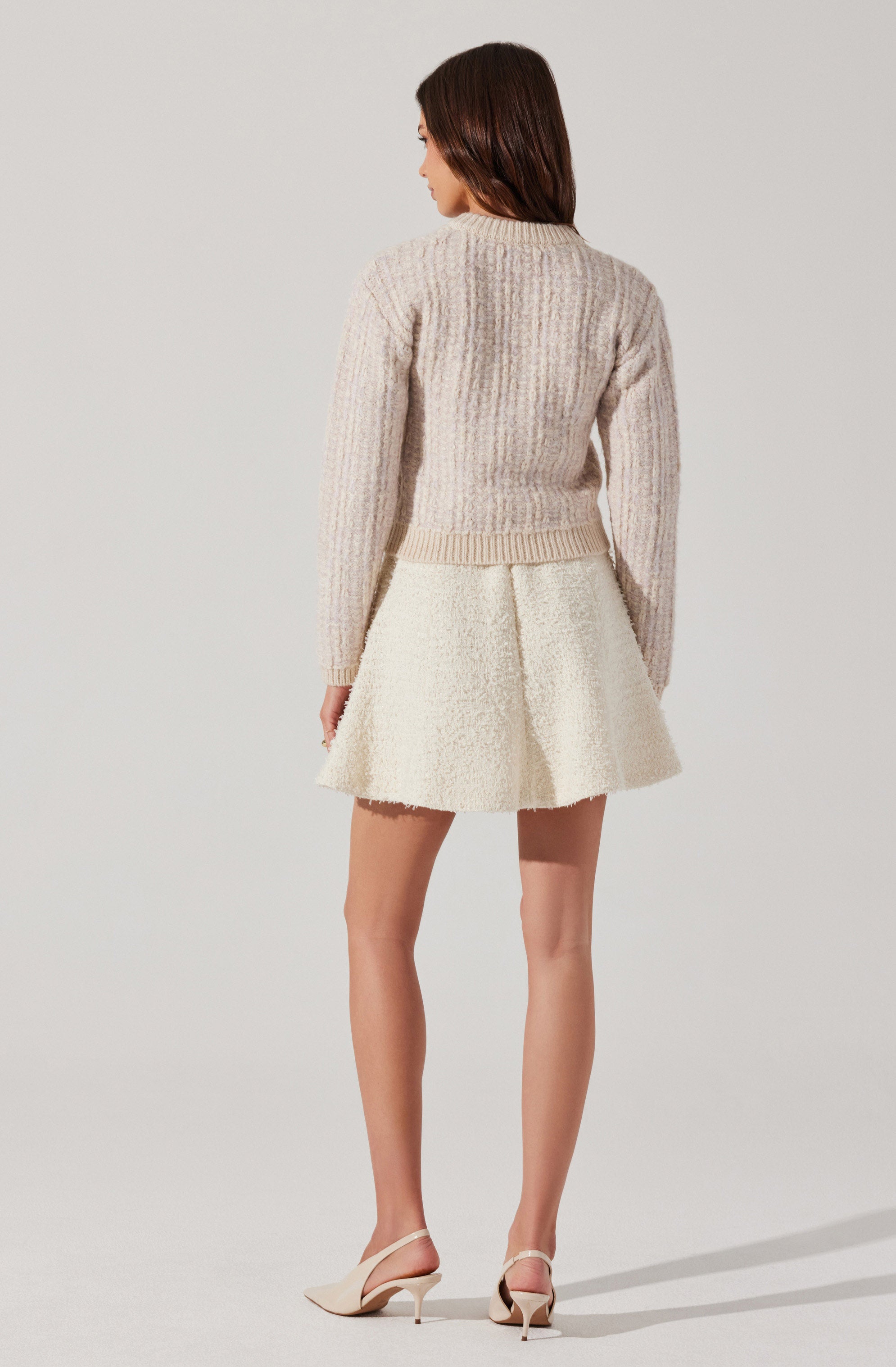 The woman is wearing a beige, textured knit sweater with a ribbed crew neckline and hem. She is also dressed in a cream-colored, flared mini skirt with a textured, bouclé-like fabric. She has on light-colored, pointed-toe high heels with ankle straps.