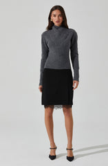 A woman is wearing a long-sleeved, high-neck, gray knit sweater with ribbed details at the cuffs and hem. She pairs it with a black knee-length skirt that features lace trim at the hem. She completes her outfit with black high-heeled shoes with ankle straps and gold earrings.