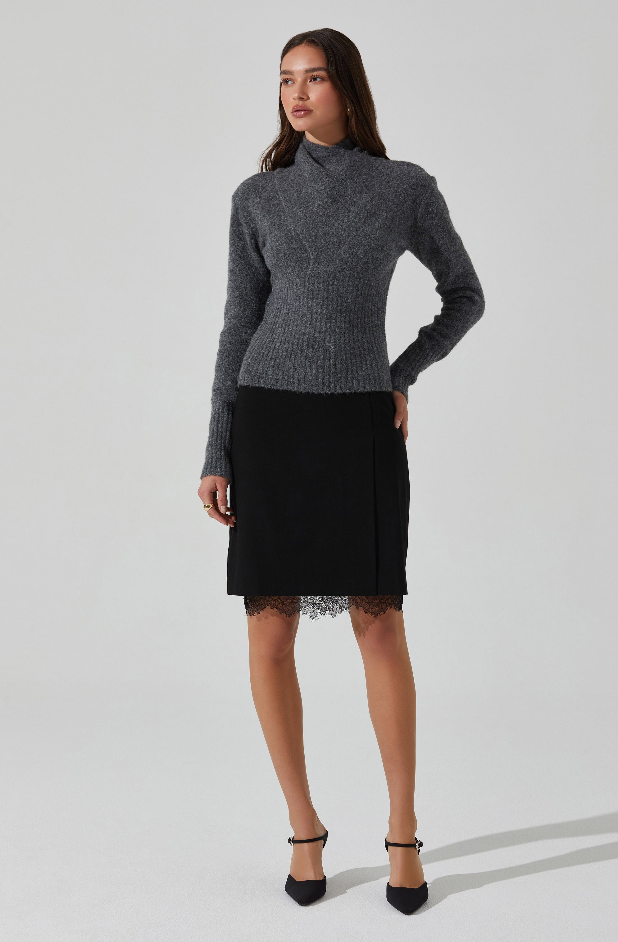 A woman is wearing a long-sleeved, high-neck, gray sweater made of a warm, textured knit fabric. She pairs it with a black skirt that falls just above the knees, featuring a lace trim at the hem. She completes her outfit with black ankle-strap heels.