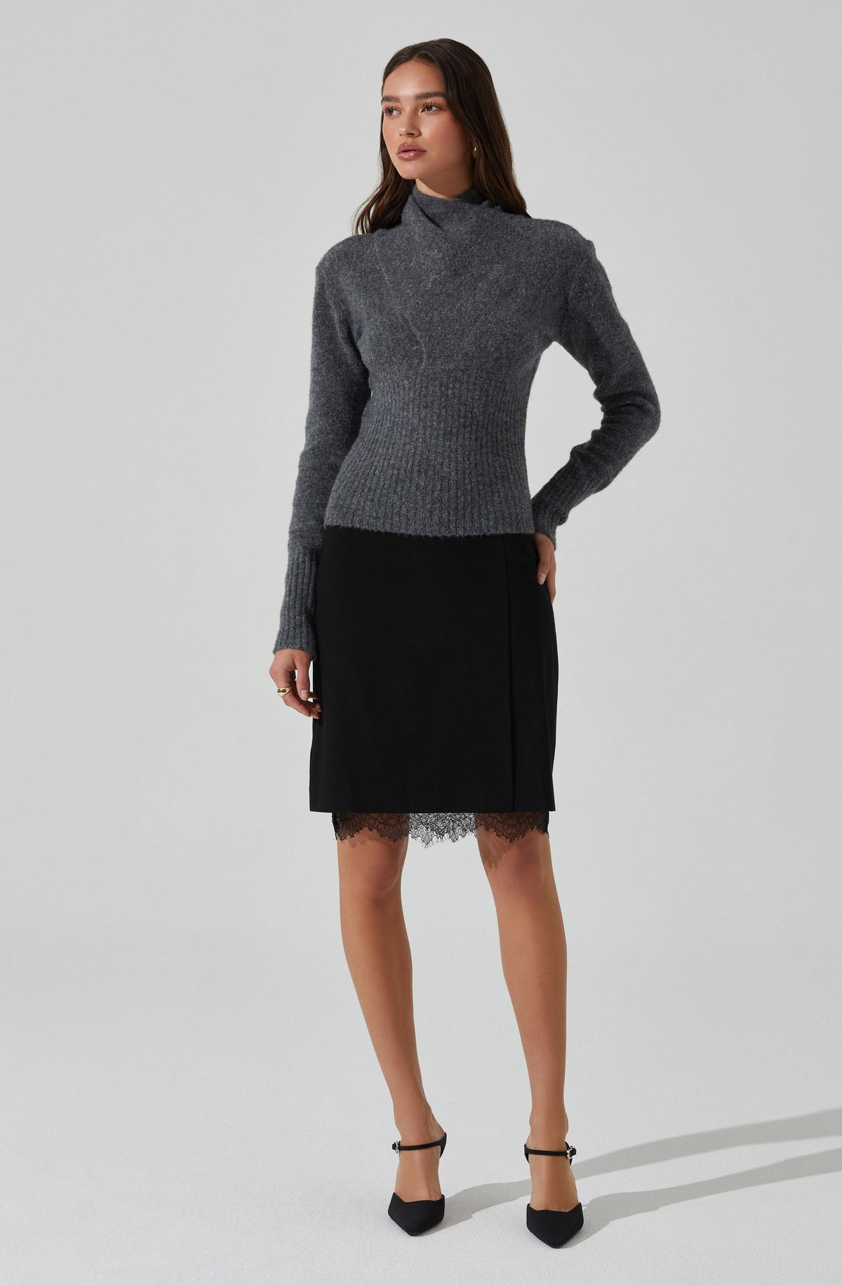 A woman is wearing a long-sleeved, high-neck, gray sweater made of a warm, textured knit fabric. She pairs it with a black skirt that falls just above the knees, featuring a lace trim at the hem. She completes her outfit with black ankle-strap heels.