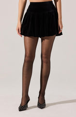 A person is wearing a black velvet mini skirt with a gathered, ruffled hem. The outfit is paired with sheer black tights and black high-heeled shoes decorated with white polka dots.