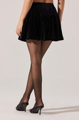 A woman is wearing a high-waisted black velvet skirt that is short and flares out in a ruffled style. She has on sheer black stockings and black high-heeled shoes decorated with small white studs. Her right hand is resting at her side, showing a simple ring on her finger.