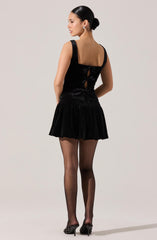 A woman is wearing a sleeveless black velvet dress with wide shoulder straps, featuring three decorative bows and cut-out details on the back. She also has on black sheer tights and black high-heeled shoes with studded embellishments. Her hair is styled in a sleek, low bun.
