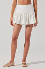 A woman is wearing a white high-waisted skirt with a gathered ruffle hem. The skirt appears to be made of a lightweight fabric, with a smooth, fitted waistband. She is also wearing beige woven flats with a T-strap design.