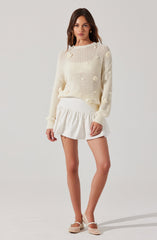 A woman is wearing a cream-colored, chunky knit sweater with a loose fit, featuring textured pom-pom details throughout. She pairs it with a white, high-waisted, ruffled mini skirt. Her accessories include gold hoop earrings, and she is wearing cream-colored, woven, low-heeled shoes with ankle straps.