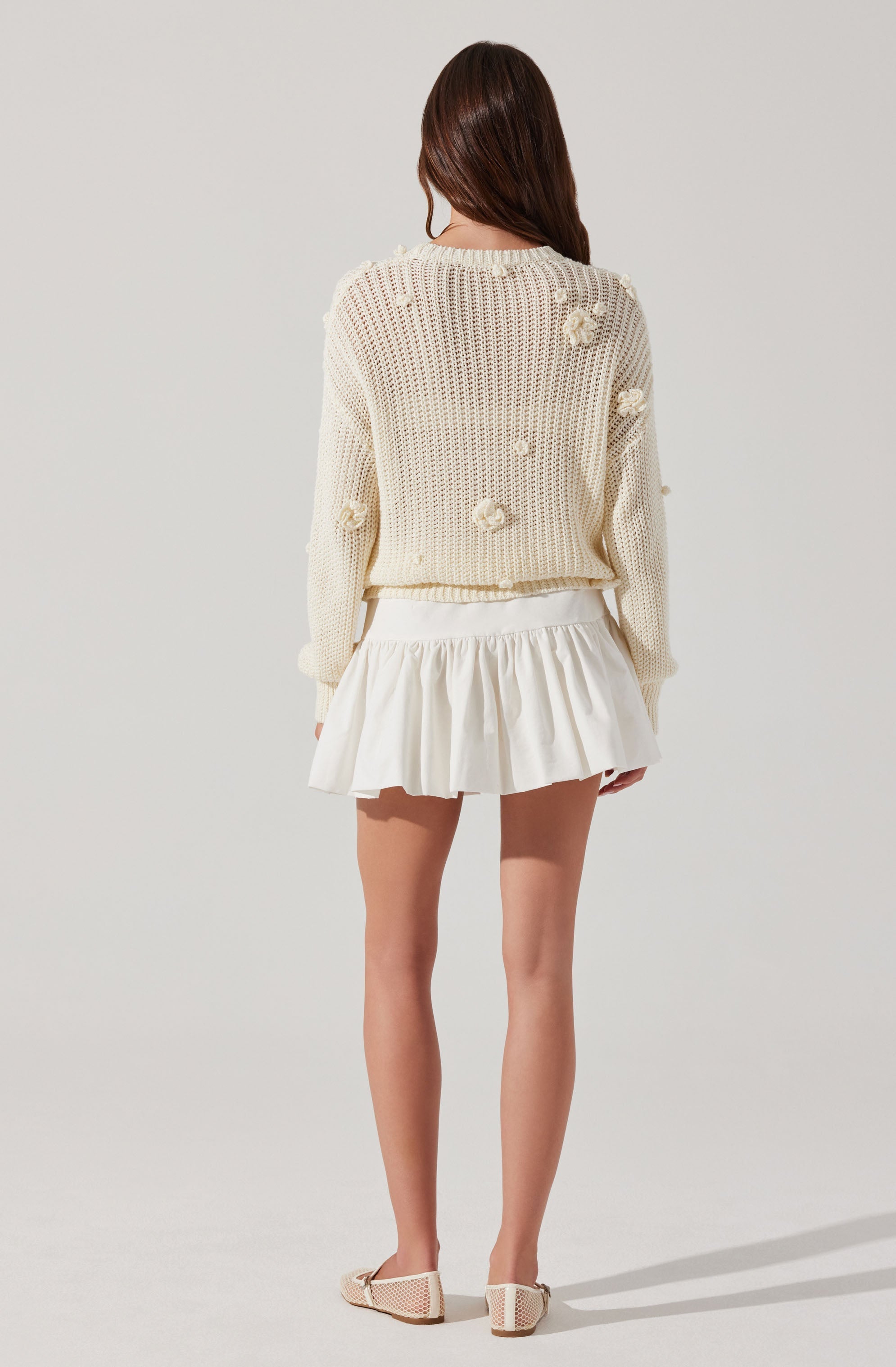 The image shows a woman wearing a cream-colored, knitted sweater with textured floral appliqués on the back and sleeves. She pairs it with a white, high-waisted, pleated mini skirt and beige, knit flats. The sweater appears loose-fitting and slightly cropped, with the floral details adding a decorative touch.